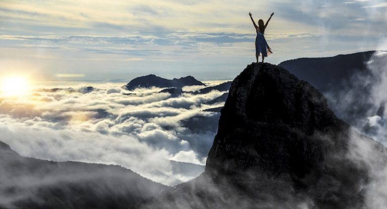 Eine Frau genießt die Sicht von einer Bergspitze über die sonnenbeleuchtete Wolkendecke.